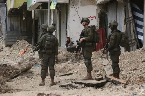 جنود إسرائيليون في منطقة مدمرة بالضفة الغربية، بينما امرأة تجلس في الخلفية. الصورة تعكس التوترات المستمرة في المنطقة.