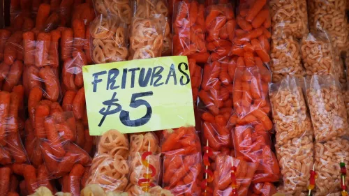 أصناف متنوعة من الوجبات الخفيفة المعبأة، تتضمن رقائق برتقالية وحلويات، مع علامة سعر مكتوبة بخط واضح، تشير إلى حظر الوجبات السريعة في المدارس المكسيكية.