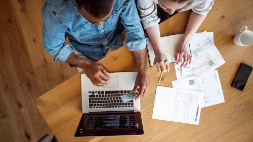 رجل وامرأة يجلسان على طاولة خشبية، يعملان على جهاز كمبيوتر محمول مع أوراق مالية وفواتير منتشرة حولهما، في سياق مناقشة الأمور المالية.