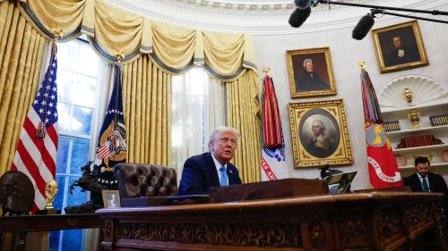 الرئيس دونالد ترامب يجلس خلف مكتبه في المكتب البيضاوي، محاطًا بالستائر الذهبية، مع العلم الأمريكي وشعار الرئاسة خلفه.