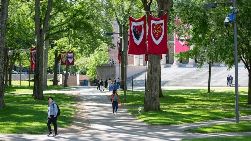 حرم جامعة هارفارد يظهر فيه طلاب يسيرون تحت لافتات الجامعة الحمراء، مع أشجار خضراء تحيط بالمكان.