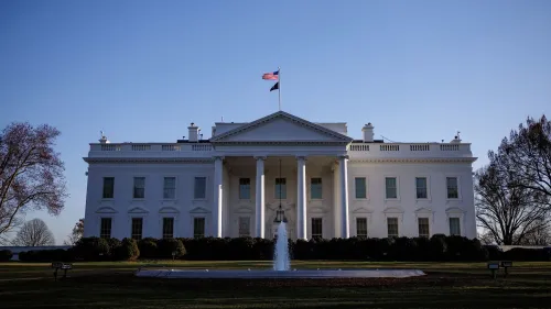 واجهة البيت الأبيض مع العلم الأمريكي يرفرف في الأعلى، تظهر حديقة أمامية ومياه نافورة، تعكس الأجواء السياسية الحالية.