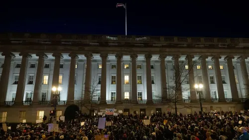 مظاهرة حاشدة أمام مبنى حكومي مضاء ليلاً، حيث يجتمع المتظاهرون حاملين لافتات تعبر عن احتجاجاتهم على سياسات إدارة ترامب.