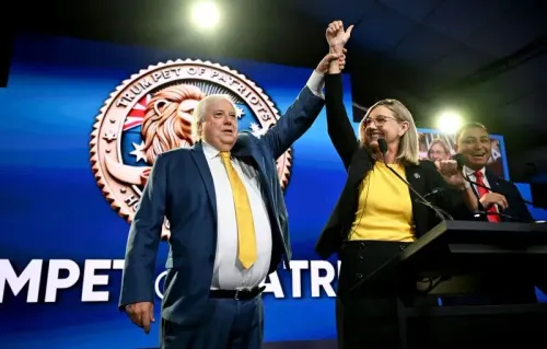 كلايف بالمر، زعيم حزب "بوق الوطنيين"، يرفع يده مع مؤيدته خلال حدث انتخابي، مع شعار مستوحى من ترامب في الخلفية.