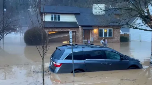 مياه الفيضانات تغمر سيارة أمام منزل، مع وجود شخصين يسيران في المياه، مما يعكس تأثير العاصفة القوية على المنطقة.