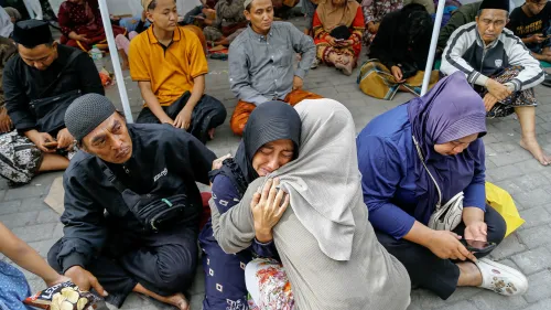 تظهر الصورة مجموعة من الأشخاص يجلسون في مكان عام، حيث تحتضن امرأة امرأة أخرى في مشهد يعكس الحزن والأمل بعد انهيار مدرسة في إندونيسيا.