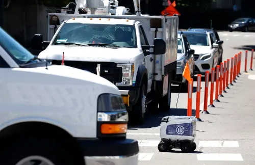 روبوت توصيل الطعام "كورتني" يتحرك بين السيارات في الشارع، مع أعمدة مرورية تحيط به، مما يعكس تحديات التنقل في المدينة.