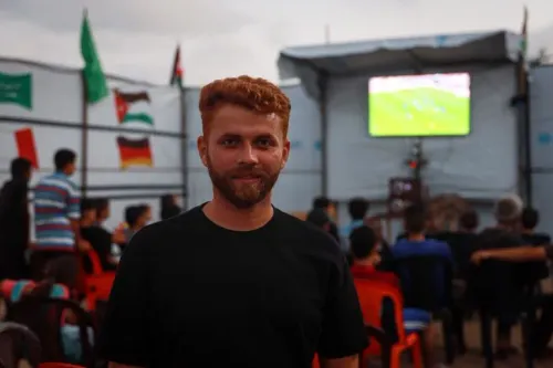 شاب مبتسم يقف أمام خيمته في استاد اليرموك بغزة، بينما تُعرض مباراة كأس العالم على شاشة خلفه، وسط أجواء من التوتر والتهجير.