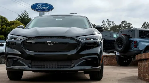 سيارة فورد موستانج ماك-إي الكهربائية معروضة في معرض سيارات، مع وجود سيارات أخرى في الخلفية، تعكس زيادة الأسعار بسبب التعريفات.