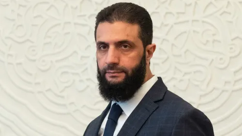 أحمد الشرع، المعروف سابقًا بأبو محمد الجولاني، في مؤتمر صحفي يعلن توليه رئاسة سوريا خلال فترة انتقالية، مع خلفية زخرفية.