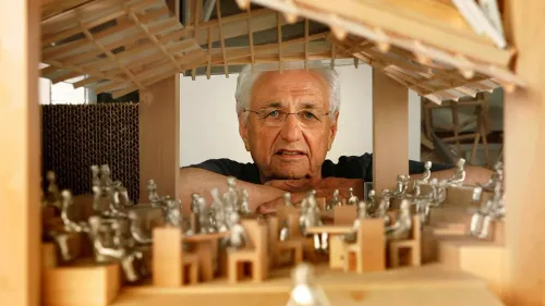 فرانك جيري، المهندس المعماري الشهير، يتأمل نموذجًا لمشروع معماري، محاطًا بتفاصيل دقيقة تعكس أسلوبه المبتكر.