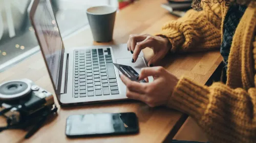 امرأة ترتدي سترة صفراء تستخدم كمبيوتر محمول للدفع عبر الإنترنت، مع وجود فنجان قهوة وكاميرا وهاتف ذكي على الطاولة.