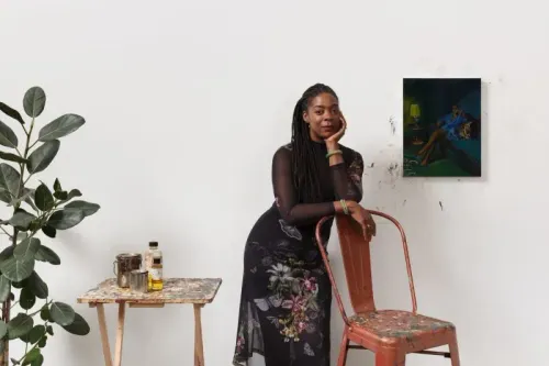 امرأة ترتدي فستانًا مزخرفًا، تجلس على كرسي بجانب طاولة، بينما تتأمل لوحة فنية على الحائط. تعكس الصورة أجواء فنية وراحة.