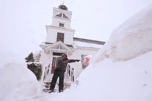 رجل يستخدم مجرفة لإزالة الثلوج من أمام مبنى خلال عاصفة شتوية قوية، مع تراكم الثلوج حوله بشكل كبير.