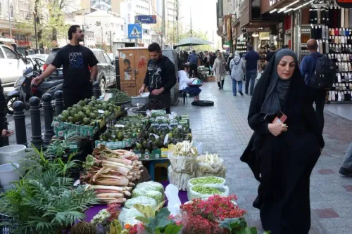امرأة ترتدي الحجاب تسير في شارع مزدحم، بينما تُعرض خضروات وفواكه طازجة على الرصيف، مما يعكس الحياة اليومية في طهران.