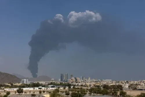 سحابة دخان كثيف تتصاعد فوق مدينة حديثة، مع خلفية للجبال والمباني، تشير إلى تأثيرات الأحداث الجارية في المنطقة.