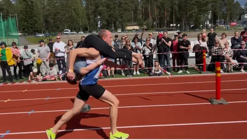 زوج يحمل زوجته على كتفيه أثناء سباق حمل الزوجات، مع جمهور يشاهد في الخلفية. تعكس الصورة أجواء المنافسة والمرح في هذا الحدث الفريد.