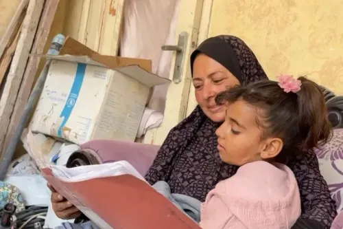 امرأة ترتدي الحجاب تجلس مع فتاة صغيرة، يتصفحان كتابًا معًا في غرفة مليئة بالركام، يعكسان لحظة من الأمل وسط المعاناة في غزة.