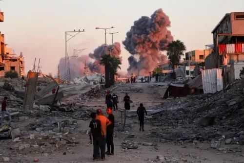 دخان يتصاعد من انفجارات في غزة، بينما يسير الناس وسط الأنقاض. الصورة تعكس آثار الحرب والدمار في المنطقة.