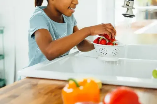 فتاة صغيرة تغسل الفراولة في مصفاة تحت صنبور ماء، مع خضروات ملونة في المقدمة، تعكس أهمية التغذية الصحية للأطفال.