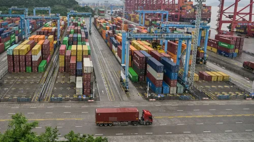 حاويات شحن ملونة مرتبة في ميناء، مع شاحنة تنقل البضائع، تعكس تأثير الحرب التجارية على الاقتصاد العالمي.