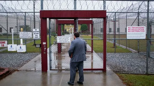 رجل يرتدي بدلة يقف أمام بوابة مغلقة لمركز احتجاز، محاط بأسلاك شائكة ولافتات تحذر من دخول الأسلحة.