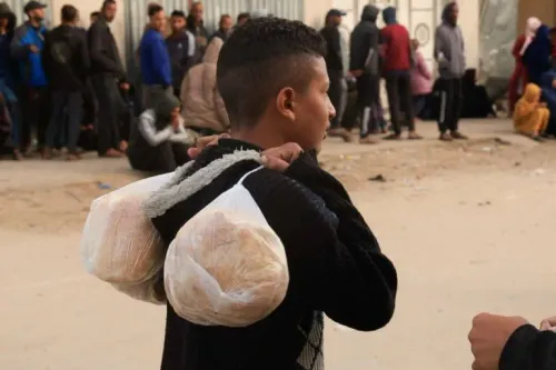 طفل يحمل خبزًا في أكياس على ظهره، منتظرًا في طابور طويل أمام مخبز في غزة، حيث يعاني السكان من نقص حاد في الدقيق بسبب القيود الإسرائيلية.