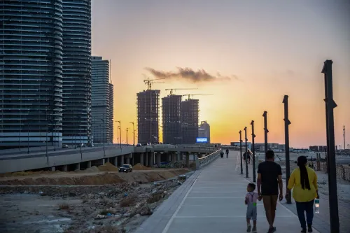 مشهد لغروب الشمس فوق ساحل مصر الشمالي، حيث يظهر تطور المشاريع العقارية مع عائلات تسير على ممشى حديث.