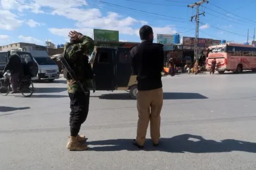 مسلح يرتدي زيًا عسكريًا يقف في الشارع مع رجل آخر، بينما تظهر حافلات ومركبات أخرى في الخلفية، في منطقة بلوشستان المضطربة.