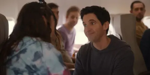 مشهد من مسلسل "Hacks" يظهر شخصية جيمي مبتسمًا أثناء حديثه مع امرأة في طائرة، مما يعكس لحظات الكوميديا والتفاعل الاجتماعي.