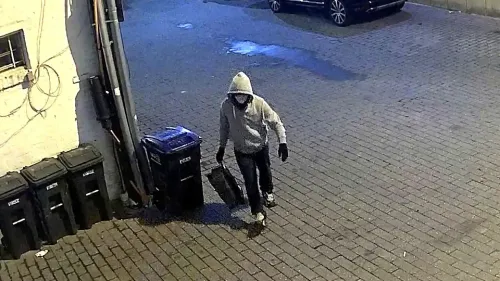 شخص يرتدي سترة رمادية وقناعًا، يحمل حقيبة في زقاق، بالقرب من حاويات القمامة، في سياق تحقيقات مكتب التحقيقات الفيدرالي حول قنابل أنبوبية.