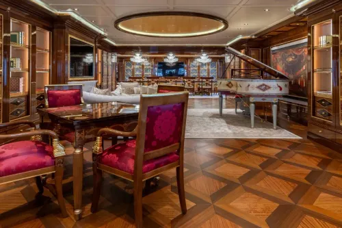 ديكور فاخر داخل يخت "أماديا" يشمل طاولة خشبية أنيقة وكراسي حمراء، مع بيانو أبيض في الخلفية، يعكس الفخامة التي يتميز بها اليخت.