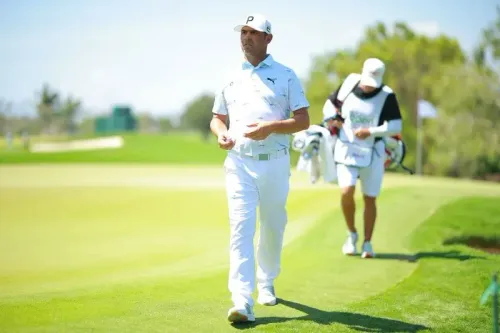 غاري وودلاند يسير على ملعب الغولف مرتديًا زيًا رياضيًا أبيض، مع وجود حامل الأكياس خلفه، مع خلفية خضراء مبهجة.