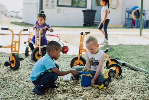 طفلان يلعبان في ساحة خارجية، حيث يقوم أحدهما بتعبئة دلو بالرمل بينما يجلس الآخر على دراجة صغيرة، مما يعكس أهمية برامج التعليم المبكر مثل Head Start.