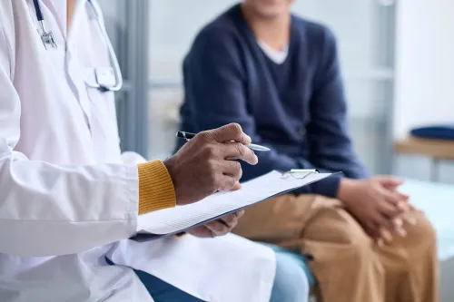 طبيب يرتدي معطفًا أبيضًا يسجل الملاحظات أثناء استشارة مع مراهق جالس بجانبه، في سياق توجيه الرعاية الصحية للشباب.