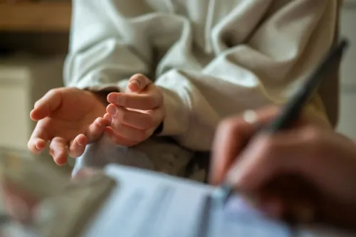 يد تظهر فيها يدي مريض تتحدثان مع طبيب أثناء زيارة عيادة، مع ملاحظات تُكتب على ورقة. تعكس الصورة أهمية التواصل في الرعاية الصحية.