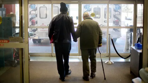 رجل مسن يمسك بيد شخص آخر أثناء مغادرته مستشفى، مع وجود سيارة إسعاف في الخلفية، مما يعكس التحديات في نظام الرعاية الصحية.