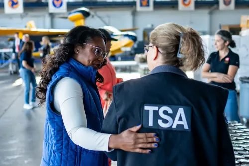 امرأة تتحدث مع موظفة من إدارة الأمن الداخلي (TSA) في موقع عمل، مع طائرات في الخلفية، مما يعكس تأثير الإغلاق الحكومي على الموظفين الفيدراليين.