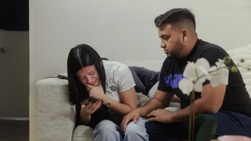 امرأة تبكي بينما تمسك بهاتفها، يجلس بجانبها رجل يواسيها، في مشهد يعكس مشاعر الحزن بسبب ترحيل أحد أفراد الأسرة.