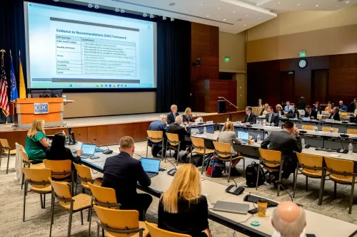 اجتماع لمجموعة من الخبراء في قاعة مركز السيطرة على الأمراض، حيث يناقشون توصيات اللقاحات مع وجود عرض تقديمي على الشاشة.