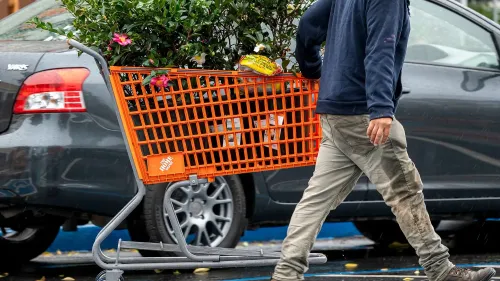 رجل يحمل عربة تسوق برتقالية من هوم ديبوت مليئة بالنباتات، بجوار سيارات في موقف للسيارات، مما يعكس نشاط المستهلكين في تحسين المنازل.
