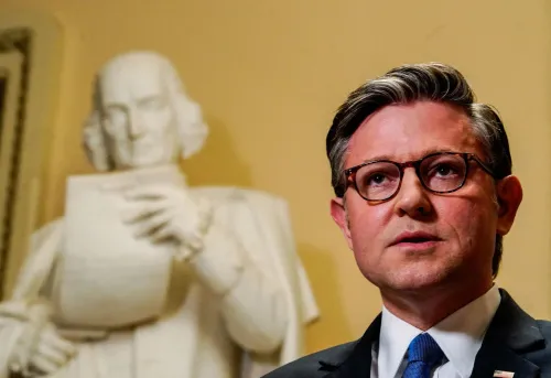 رجل يرتدي نظارات وقميصاً رسمياً يتحدث بجدية، بينما تظهر خلفه تمثال لرجال تاريخيين يحملون وثيقة، في سياق مناقشات سياسية.