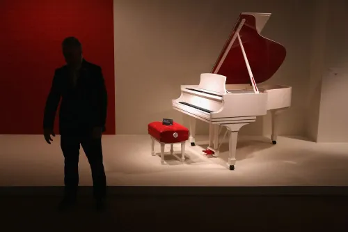 بيانو أبيض أنيق مع غطاء أحمر، بجانبه مقعد مخصص، يظهر في معرض. يرمز إلى الحرفية العالية في صناعة البيانو الأمريكية.