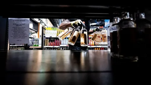 يد تظهر شخصًا يرتدي قفازات وهو يحمل زجاجات مشروبات كحولية من رف في متجر، مع وجود لافتة توضح تأثير الرسوم الجمركية على الواردات الأمريكية.