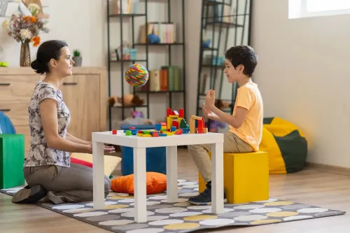 امرأة وطفل يلعبان معًا على طاولة، حيث يقوم الطفل ببناء هياكل باستخدام مكعبات ملونة، مما يعكس تفاعلًا إيجابيًا وداعمًا.