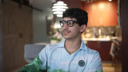 شاب يرتدي نظارات وقميص مزخرف يجلس في غرفة معيشة، مع خلفية تحتوي على مطبخ وأضواء خافتة، يعبر عن حماسه لحفلات باد باني في بورتوريكو.