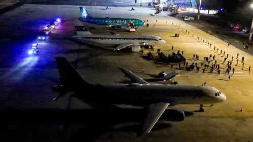 صورة تظهر عدة طائرات في مطار ليلاً، مع وجود رجال أمن ومجموعات من الأشخاص في الخلفية، تعكس مشهد الترحيل المثير للجدل.