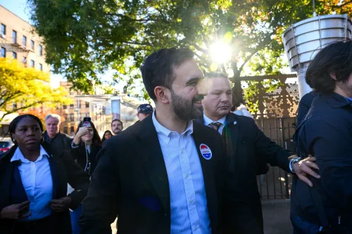 مرشح عمدة نيويورك زهران ممداني يتحدث مع مؤيديه في شارع مزدحم، محاطًا بأفراد من حملته الانتخابية، مع شمس ساطعة في الخلفية.