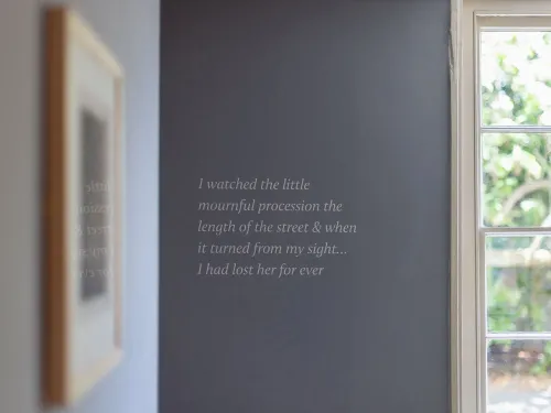 نص على الجدار داخل منزل جين أوستن يعبر عن مشاعر الفقد، مع نافذة تطل على الحديقة، مما يبرز اللحظات الأخيرة في حياتها.