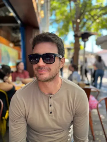 رجل يرتدي نظارات شمسية يجلس في مقهى في الهواء الطلق، محاط بأشخاص آخرين، يعكس أجواء الحياة اليومية والتفاعل الاجتماعي.